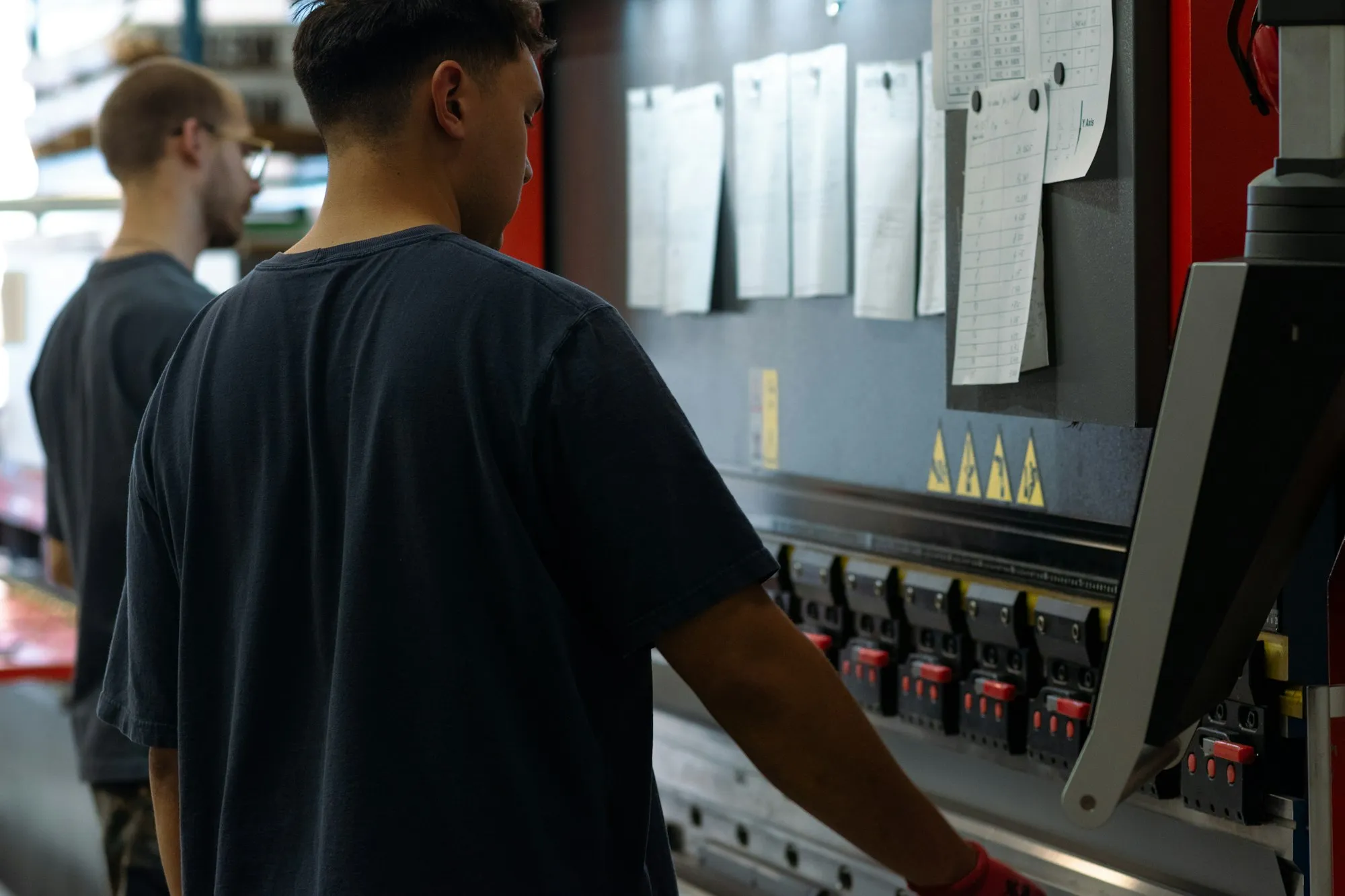 Deux opérateurs devant une presse plieuse, avec des fiches de travail papier accrochées sur le bâti de la machine.