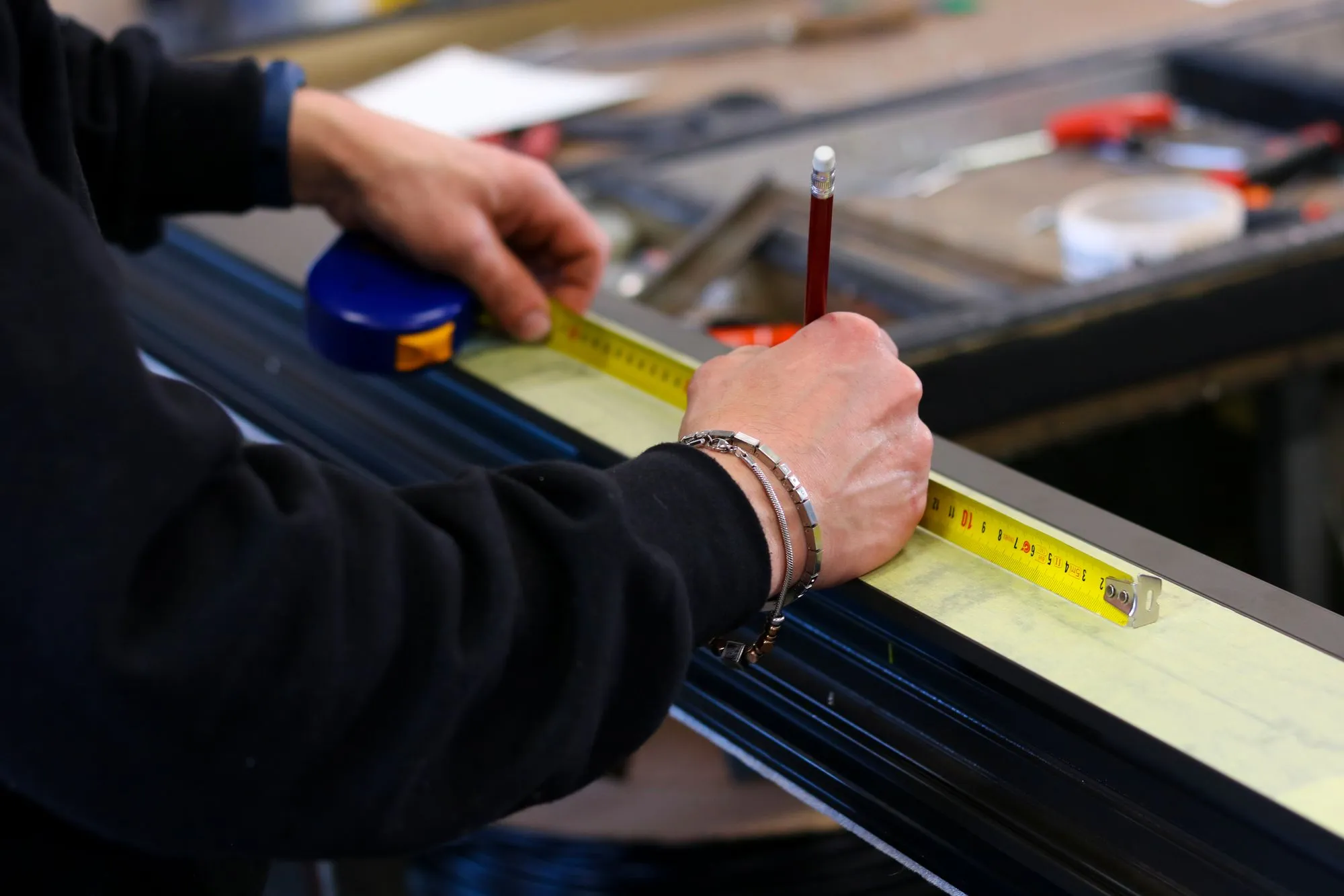 Gros plan sur les mains d’un menuisier qui mesure un profilé aluminium au mètre ruban et trace un repère au crayon.