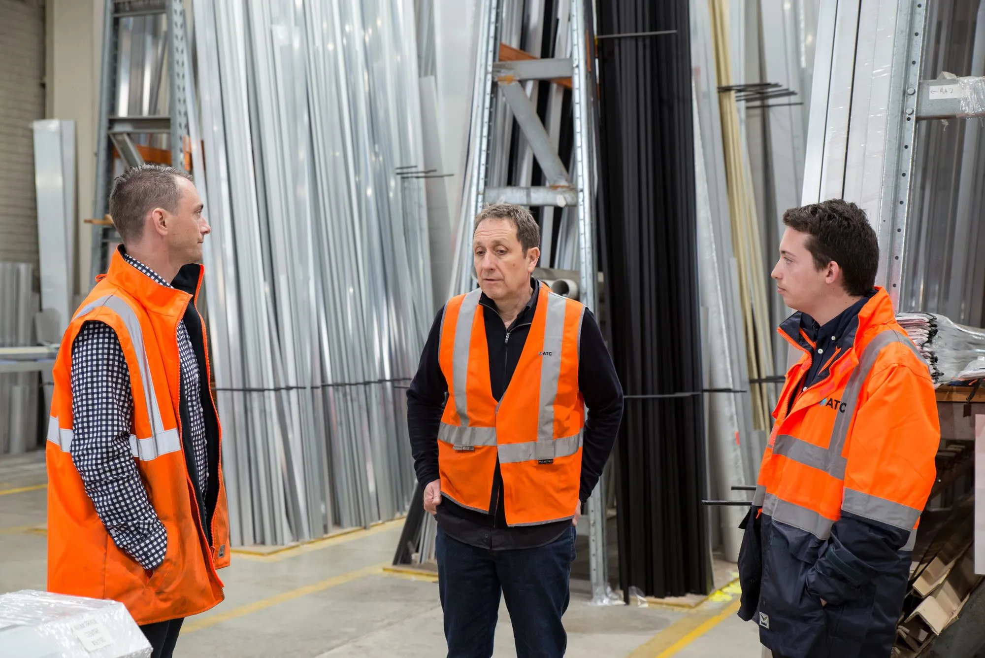 Trois hommes en gilet orange discutent debout dans un stock de barres et de profilés métalliques.